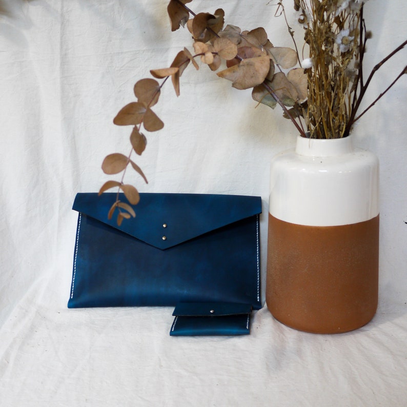 May include: A blue leather envelope clutch with a smaller matching wallet. The clutch has a flap closure and a white stitch detail around the edges. The wallet has a simple design with a flap closure.
