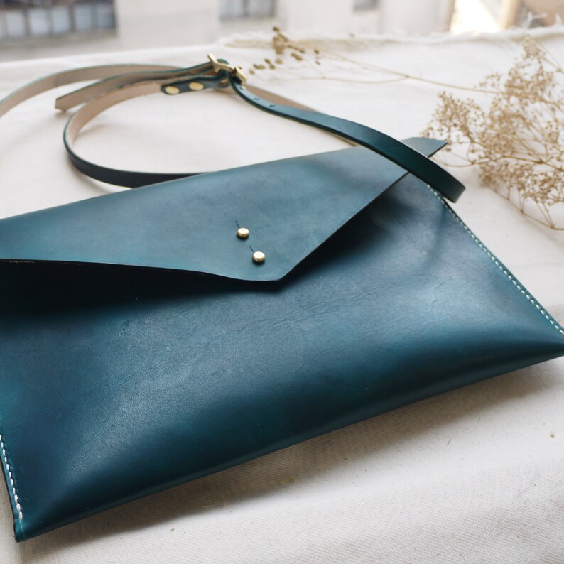 Op de afbeelding: Een teal leren envelop clutch met een lange band. De clutch heeft twee gouden metalen klinknagels op de voorste klep.