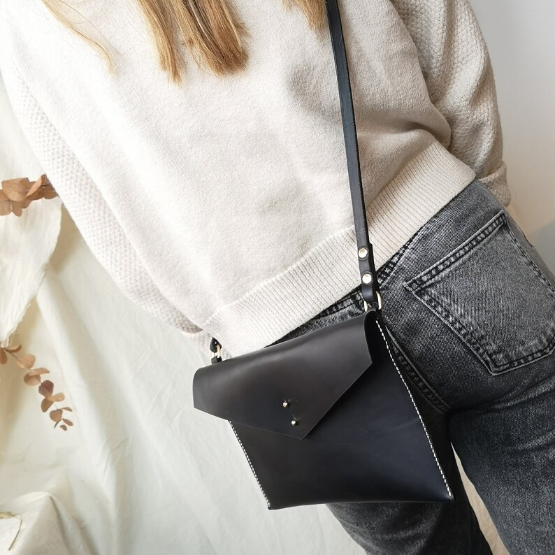Op de afbeelding: Een zwarte leren crossbody tas in envelopstijl met een lange band. De tas heeft een klep sluiting en wordt gedragen over een witte trui en een donkere spijkerbroek.