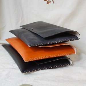May include: Three leather laptop sleeves stacked on top of each other. The top sleeve is dark blue, the middle sleeve is orange, and the bottom sleeve is black. All three sleeves have decorative stitching around the edges.