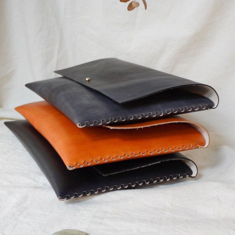 May include: Three leather laptop sleeves stacked on top of each other. The top sleeve is dark blue, the middle sleeve is orange, and the bottom sleeve is black. All three sleeves have decorative stitching around the edges.