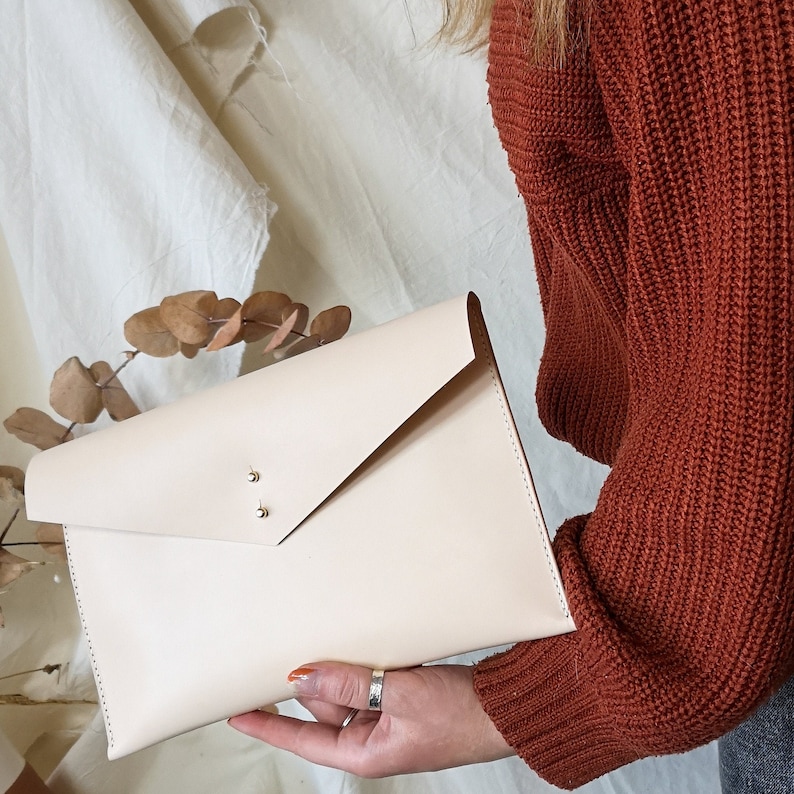 May include: A light beige leather clutch with a flap closure and gold accents. The clutch is held in a hand wearing a gold ring.