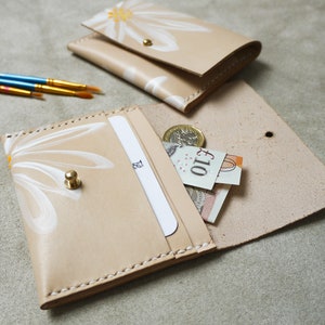 May include: A beige leather wallet with a white daisy design and a gold button closure. The wallet is open and has a card slot and a cash pocket. There is a &pound;10 note and a coin inside the wallet.