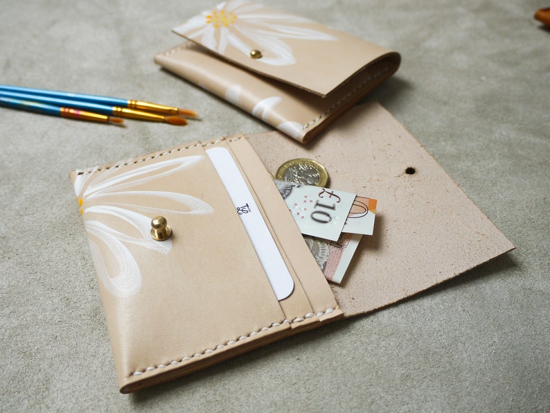 May include: A beige leather wallet with a white daisy design and a gold button closure. The wallet is open and has a card slot and a cash pocket. There is a &pound;10 note and a coin inside the wallet.