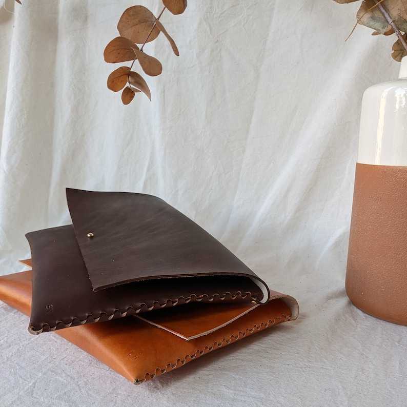 May include: Three leather envelopes stacked on top of each other. The top envelope is dark brown, the middle envelope is light brown, and the bottom envelope is a lighter shade of brown. All three envelopes have decorative stitching around the edges.