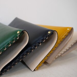 May include: Four leather wallets in different colors, including green, black, yellow, and cream. The wallets are folded and stacked on top of each other. Each wallet has a decorative stitched edge and a brass rivet.