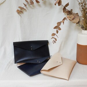 May include: Three leather envelopes in different colors: black, navy blue, and beige. The envelopes are stacked on top of each other, with the black envelope on top.