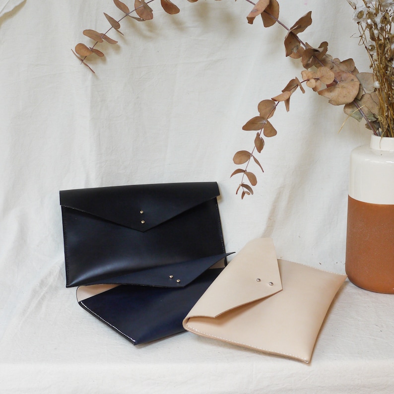May include: Three leather envelopes in different colors: black, navy blue, and beige. The envelopes are stacked on top of each other, with the black envelope on top.