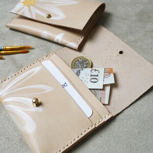 May include: A beige leather wallet with a white daisy design. The wallet is open and has a card slot and a cash compartment. There is a &pound;10 note and a coin inside the wallet.