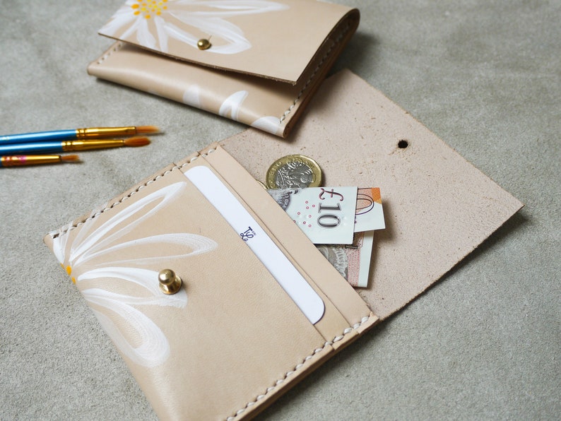 May include: A beige leather wallet with a white daisy design. The wallet is open and has a card slot and a cash compartment. There is a &pound;10 note and a coin inside the wallet.