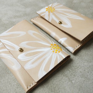 May include: Two beige leather wallets with white daisy designs and yellow centers. The wallets have brass snaps.
