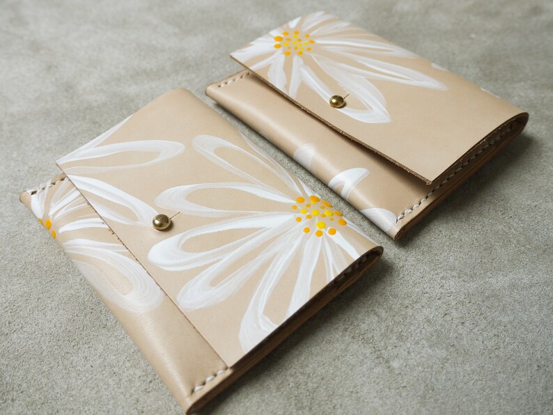 May include: Two beige leather wallets with white daisy designs and yellow centers. The wallets have brass snaps.