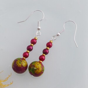 May include: A pair of dangle earrings with a burgundy and gold floral design. The earrings feature a large burgundy bead with a gold floral pattern and smaller burgundy beads with gold accents.