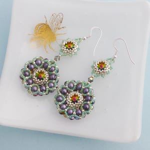 May include: Handmade beaded earrings with a floral design. The earrings feature a central iridescent bead surrounded by purple and green beads. The earrings are on a white surface with a gold bee illustration.