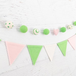 May include: A green and pink felt garland with flags and balls. The flags are alternating pink and green, and the balls are green with white polka dots, green with white swirls, and white with green polka dots. There is also a pink felt heart.