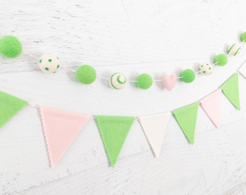 May include: A green and pink felt garland with flags and balls. The flags are alternating pink and green, and the balls are green with white polka dots, green with white swirls, and white with green polka dots. There is also a pink felt heart.