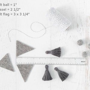 May include: A close-up of craft supplies for making a felt garland. The supplies include gray felt triangles, gray felt balls, gray tassels, and white twine. The text "felt ball = 1", "tassel = 2 1/2", and "felt flag = 3 x 3 1/4" is written on a white surface.