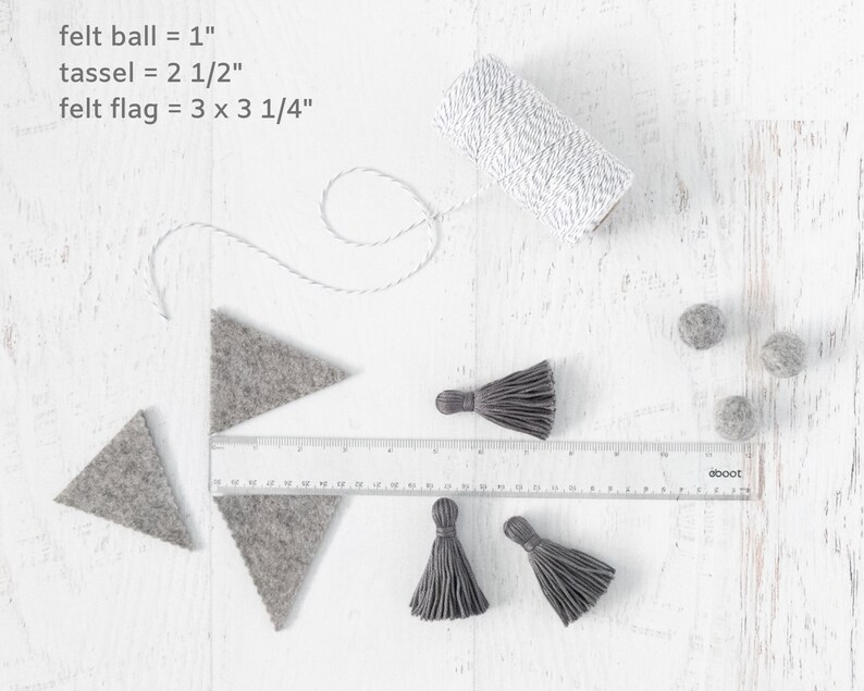 May include: A close-up of craft supplies for making a felt garland. The supplies include gray felt triangles, gray felt balls, gray tassels, and white twine. The text "felt ball = 1", "tassel = 2 1/2", and "felt flag = 3 x 3 1/4" is written on a white surface.