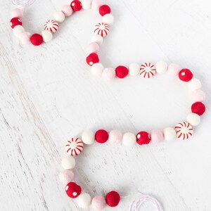 May include: A white string garland with red and pink felt balls and peppermint candy ornaments. The garland is on a white wooden surface.