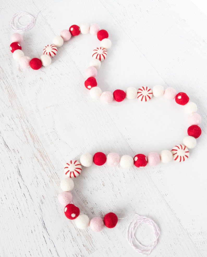 May include: A white string garland with red and pink felt balls and peppermint candy ornaments. The garland is on a white wooden surface.