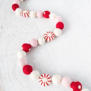 May include: A festive garland made of felt balls in red, white, and pink. The garland features alternating colors and a peppermint candy design.