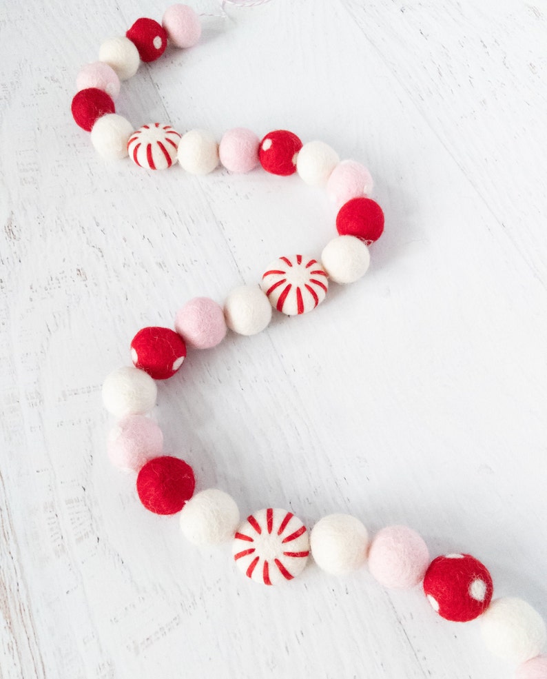May include: A festive garland made of felt balls in red, white, and pink. The garland features alternating colors and a peppermint candy design.