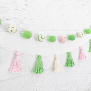 May include: A green and pink felt ball garland with white tassels. The garland is strung on a white string and has a variety of different shapes and sizes of felt balls.