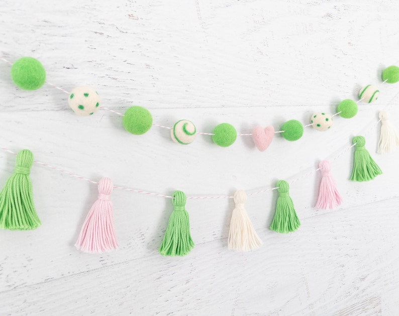 May include: A green and pink felt ball garland with white tassels. The garland is strung on a white string and has a variety of different shapes and sizes of felt balls.
