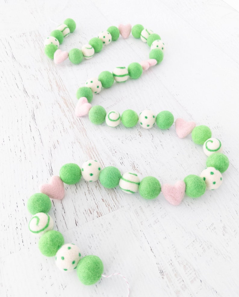 May include: A green and white felt ball garland with pink felt hearts. The garland is made of felt balls with different patterns, including polka dots, stripes, and swirls.