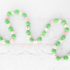 May include: A green and pink felt ball garland with white polka dots and swirls. The garland is strung together with a white thread and is shaped into a heart.