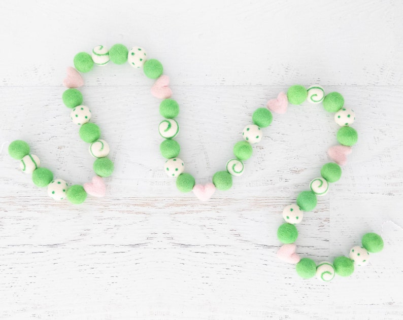 May include: A green and pink felt ball garland with white polka dots and swirls. The garland is strung together with a white thread and is shaped into a heart.
