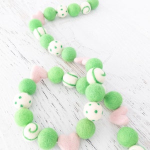 May include: A green and pink felt ball garland with white polka dots, stripes, and swirls. The garland is made of felt balls and hearts.