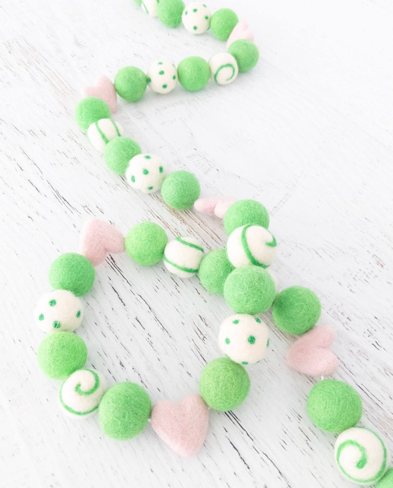 May include: A green and pink felt ball garland with white polka dots, stripes, and swirls. The garland is made of felt balls and hearts.