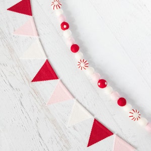 May include: A white background with a red and white felt garland and a red, white, and pink felt banner. The garland is made of pom-poms and candy cane shapes. The banner is made of triangles.