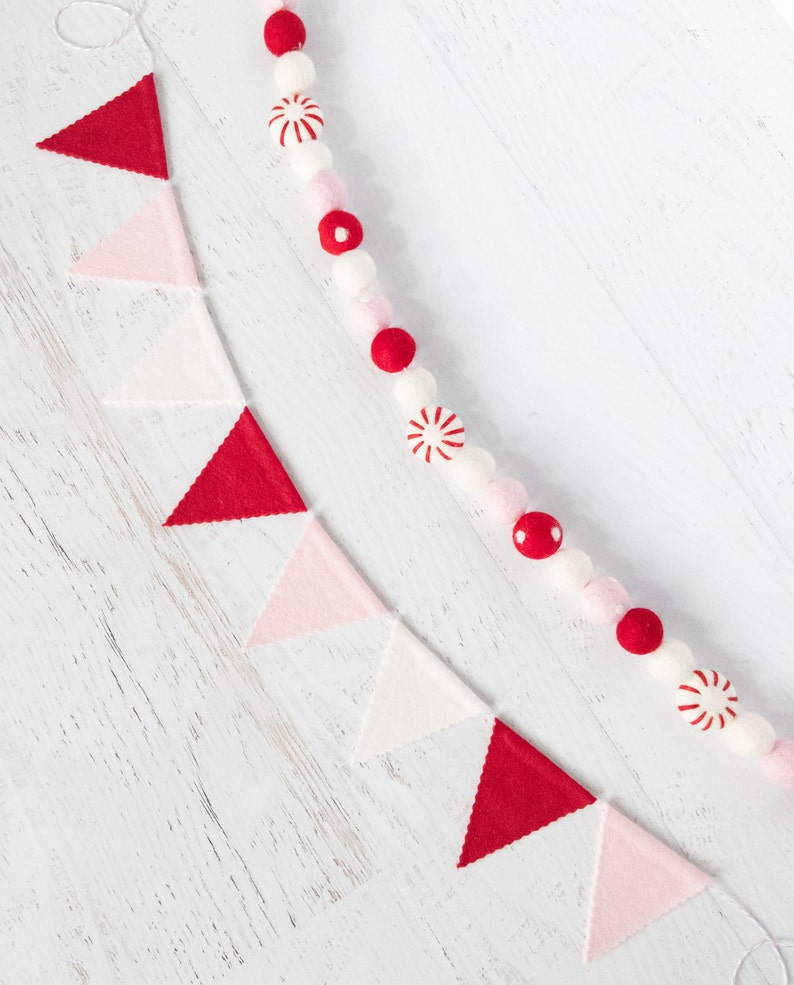 May include: A white background with a red and white felt garland and a red, white, and pink felt banner. The garland is made of pom-poms and candy cane shapes. The banner is made of triangles.