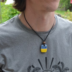 May include: A person wearing a gray t-shirt with a palm tree graphic and a necklace with a blue and yellow square pendant.