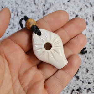 Op de afbeelding: Witte keramische hanger met een zonontwerp en een gat in het midden. De hanger is aan een zwart koord met een houten kraal.