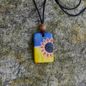 May include: A rectangular pendant necklace with a sunflower design. The pendant is divided into blue and yellow sections, with a sunflower in the center. The sunflower has a black center and red-orange petals. The necklace has a black cord and a wooden bead.