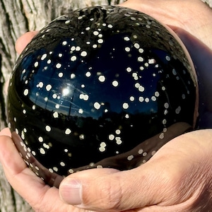 May include: A black, spherical decorative object with white speckles, held in cupped hands. The glossy surface reflects light, creating highlights. The sphere's design resembles a starry night sky, adding a touch of celestial charm.