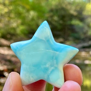 May include: A light blue star-shaped crystal. The crystal has a translucent appearance with a mix of light blue and white colors. The star is held in a person's hand, with a blurred green background.