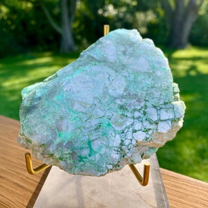 May include: A large, green and white, irregularly shaped, polished, natural stone on a gold metal stand. The stone is on a white acrylic base.