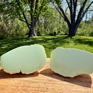 May include: Two large, light green, smooth, polished, natural stone slabs. The slabs are irregularly shaped and have a rough, natural edge. The slabs are sitting on a wooden surface.