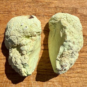 May include: Two irregularly shaped green stones with a rough, textured surface. The stones are light green in color and have a slightly yellowish hue. The stones are placed side-by-side on a wooden surface.