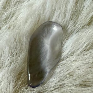 May include: A smooth, translucent, gray-colored, oval-shaped stone with a white, cloudy pattern inside. The stone is resting on a white, fluffy surface.
