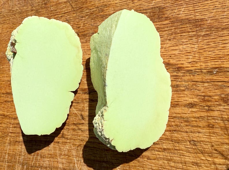 May include: Two pieces of light green polished stone, shaped like a heart, on a wooden surface.