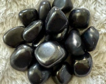 May include: A collection of polished, black hematite stones. The stones have a smooth, reflective surface and vary in size and shape. They are arranged in a pile on a soft, light-colored, textured surface.