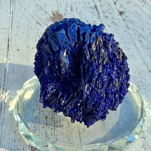 May include: A deep blue azurite mineral specimen with a crystalline structure, resting on a clear, faceted glass dish. The mineral's color is a rich, saturated blue, with hints of silver. The dish is clear and reflects light.