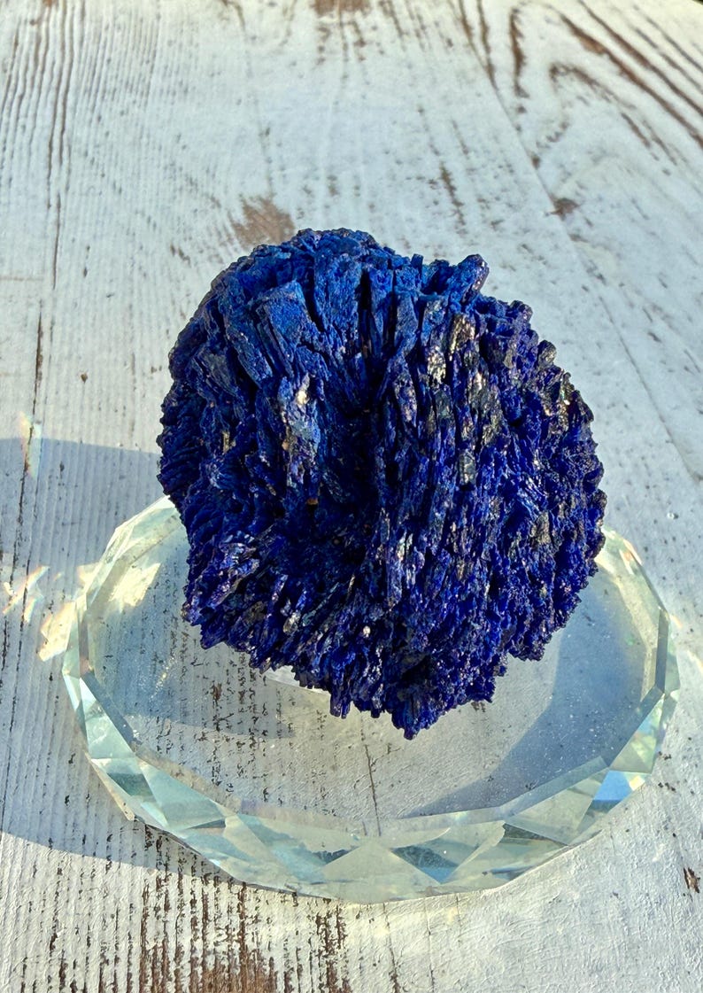 May include: A deep blue azurite mineral specimen with a crystalline structure, resting on a clear, faceted glass dish. The mineral's color is a rich, saturated blue, with hints of silver. The dish is clear and reflects light.