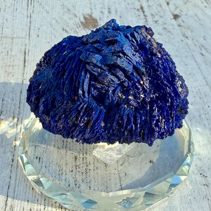 May include: A vibrant blue azurite mineral specimen with a textured, crystalline surface. The mineral sits atop a clear, faceted glass display stand, reflecting light. The background is a weathered, white wooden surface.