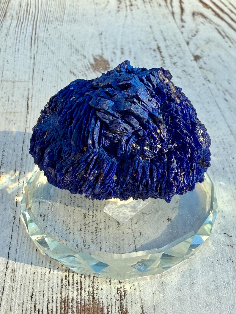 May include: A vibrant blue azurite mineral specimen with a textured, crystalline surface. The mineral sits atop a clear, faceted glass display stand, reflecting light. The background is a weathered, white wooden surface.